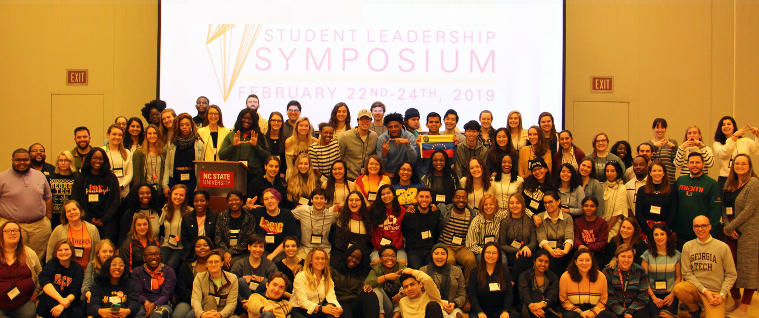 Large group of students in front of Student Leadership Symposium slide on a projector screen